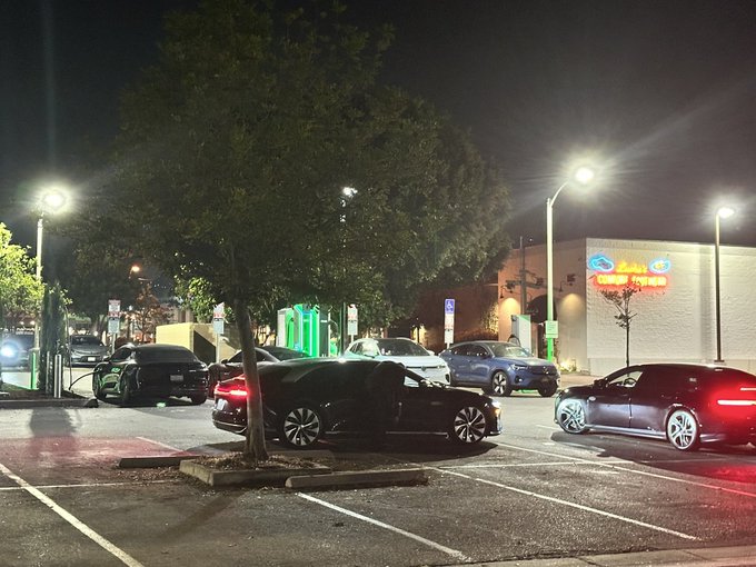A nighttime parking lot scene with several cars, a large tree, bright streetlights, and a building with a neon sign in the background.