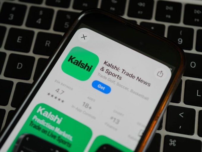 The image shows the Kalshi app on a smartphone, with the app's listing "Kalshi: Trade News & Sports" visible alongside its 4.7 star rating. The app sits on a laptop keyboard, visually connecting to the post about Baltimore officials alleging that Kalshi and Polymarket are operating unlicensed sportsbooks under the guise of "prediction markets."