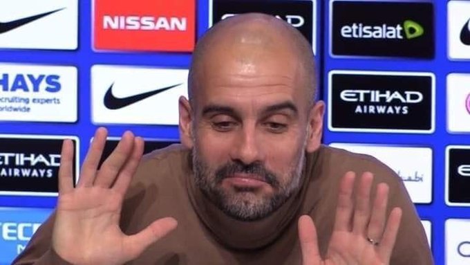 A bald man with a beard, wearing a brown t-shirt, gestures with both hands raised during a press conference, with sponsor logos including Nissan, Nike, Etisalat, and Etihad Airways visible on the backdrop behind him.
