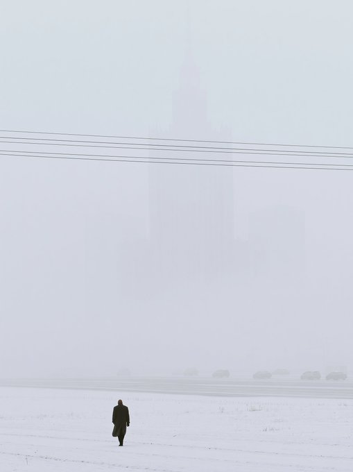 A bleak, foggy winter scene with a lone person in a dark coat walking across a snowy field toward a barely visible skyscraper looming in the mist, with power lines stretching across the gray sky.