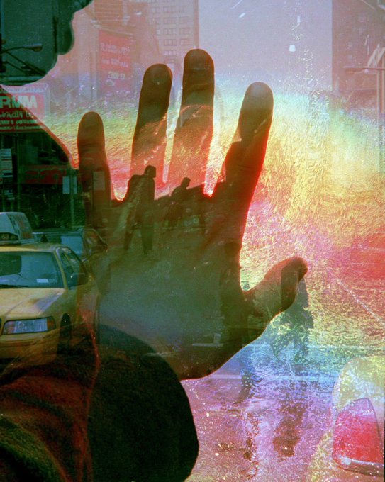 An artistic double-exposure photo showing a large hand silhouette overlaid on a colorful, textured urban street scene with taxis and pedestrians.