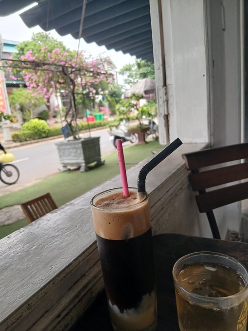 A café window view showing a tall iced coffee with a pink straw and a shorter iced drink on a wooden counter, looking out onto a street with potted bougainvillea and a passing motorbike.
