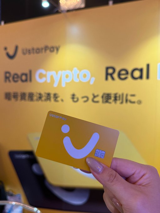 A hand holds up a yellow UstarPay debit card in front of a yellow WebX backdrop featuring the "UstarPay – Real Crypto, Real..." slogan and Japanese text, serving as the visual riddle for the quiz question asking how many cards appear in the backdrop.

Citation:
- UstarPay logo and "Real Crypto, Real..." text visible on yellow backdrop
- Japanese text: 暗号資産決済を、もっと便利に。(Crypto payments, more convenient.)