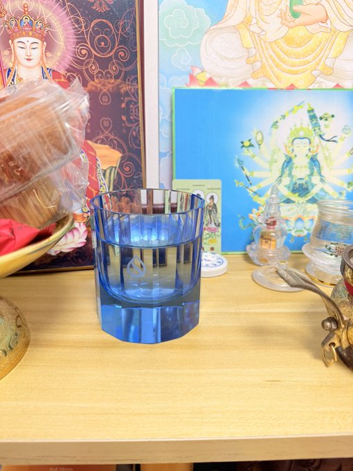 A heavy blue ribbed glass cup, a giveaway from Sui Network's April event, sits on a wooden altar surrounded by Buddhist imagery and figurines, now repurposed as a daily ceremonial vessel.