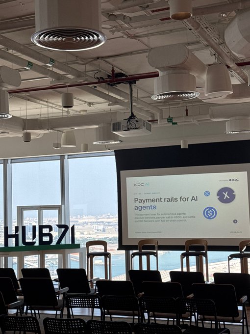 Empty event room with rows of black chairs facing a projector screen displaying a presentation titled "Payment rails for AI agents" powered by USDC on the x402 protocol, with "HUB71" signage by the window.