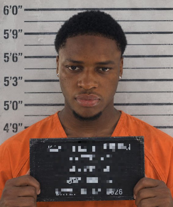 A young man in an orange prison jumpsuit holds a blurred booking placard in front of a height chart measuring background.