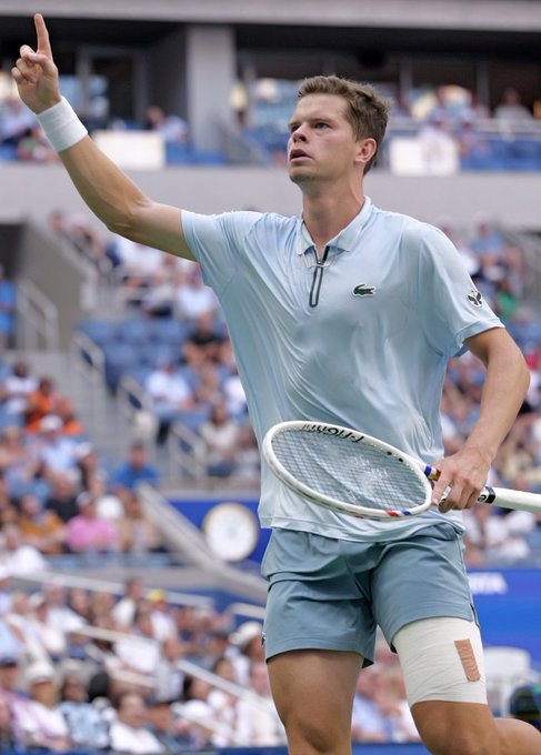 A male tennis player in a light blue Lacoste shirt raises his index finger triumphantly while holding his racket during a match, likely at the US Open given the stadium setting, illustrating the underdog betting scenario highlighted in the post.
