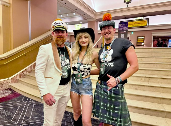 Three people pose together in costume; the man on the left wears a white captain's uniform, the woman in the middle wears a black hat and a cow-print top with denim shorts, and the man on the right wears a kilt and a Tam o' shanter.