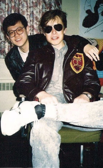 A vintage photograph shows two young men sitting together, with one wearing sunglasses, a striped shirt, and a black leather jacket featuring a patch.