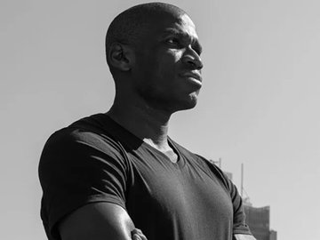A black-and-white portrait photograph of a man in a dark t-shirt shown in profile, with city buildings visible in the background.