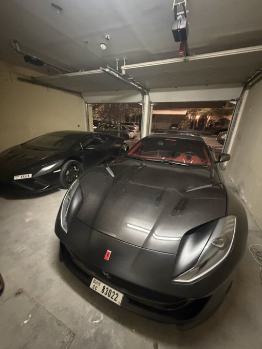 A matte black Ferrari with a red interior and license plate "CC 83022" parked in a garage next to a dark Lamborghini.
