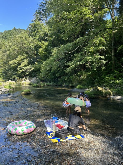 A family enjoys a day of river tubing in a lush forest setting, with two children carrying a float into the water while another person sits on a picnic mat beside an inner tube. The "Charter!" caption suggests a summer adventure or recreational outing, fitting perfectly with this scene of leisurely water play.