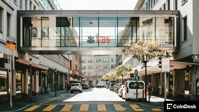 A street view photograph of a UBS bank building with an enclosed glass pedestrian bridge crossing over the road, marked with a 'CoinDesk' watermark.
