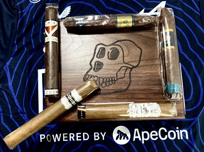 An arrangement of premium cigars displayed on a wooden board engraved with a stylized skull design, resting on a dark blue sports jersey featuring 'POWERED BY ApeCoin' text at the bottom.