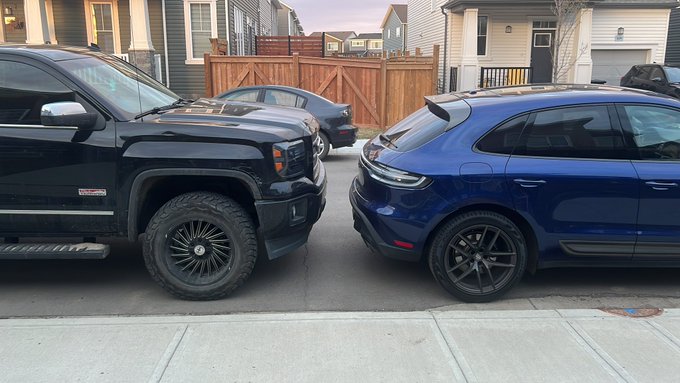 A black GMC Sierra pickup truck faces off against a blue Porsche Macan in a suburban driveway, humorously visualizing the wealth disparity highlighted by the post's joke about "majors" versus "meme coins." The contrasting vehicles—one a practical workhorse, the other a luxury sports SUV—symbolize the financial divide between traditional investments and speculative crypto.