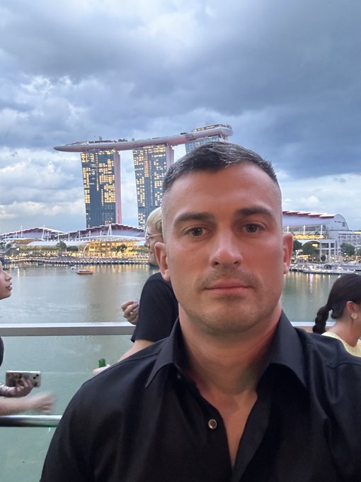 A man in a black shirt poses for a selfie at Marina Bay Sands in Singapore, with the iconic three-tower hotel and its rooftop SkyPark visible in the background under a cloudy evening sky. The setting aligns with the post about TOKEN2049 Singapore, capturing the event's host city destination.