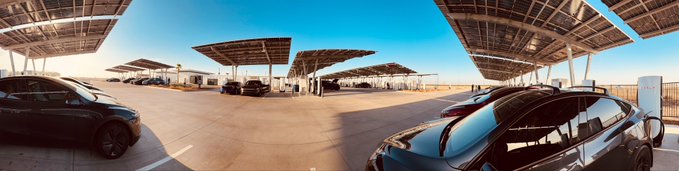 A panoramic view of an EV charging station with solar panel canopies, showing several cars parked at charging stalls under a clear blue sky.