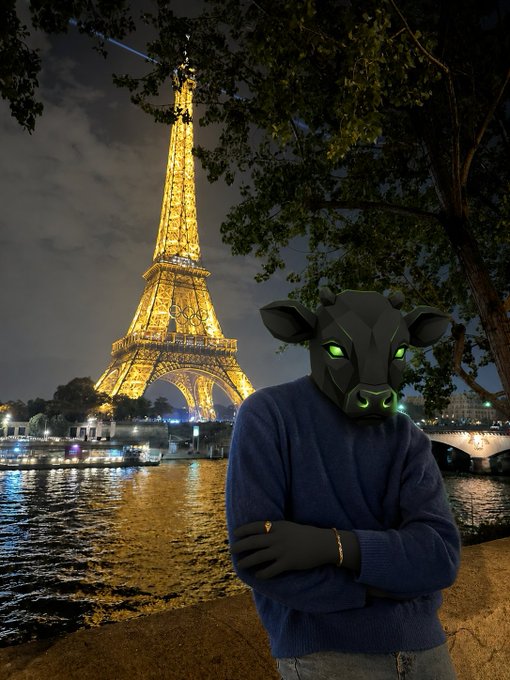 A surreal photo featuring a person wearing a black bull mask with glowing green eyes and a blue sweater, standing with arms crossed in front of the illuminated Eiffel Tower at night in Paris.