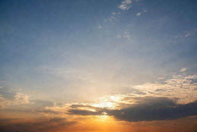 A serene sunset sky with the sun breaking through scattered clouds, casting golden rays across a gradient of blue and orange hues.