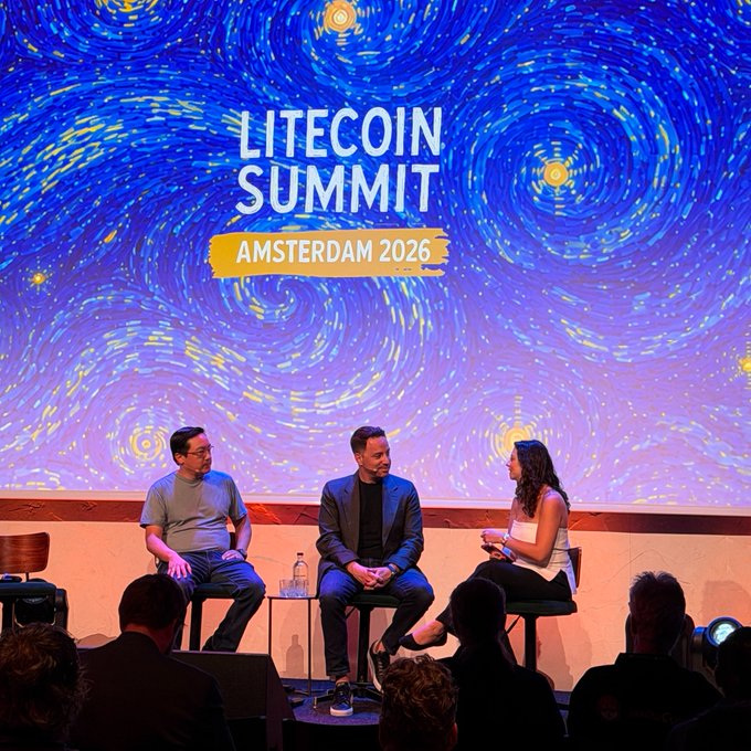 The image shows three men sitting at a table with a microphone, likely participating in a panel discussion or interview during the Litecoin Summit event held in Amsterdam, Netherlands. The event's organizers are promoting Litecoin as an investment opportunity for institutional investors.