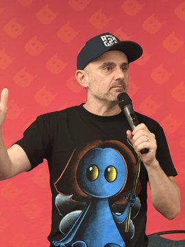 A man wearing a dark baseball cap and a black t-shirt featuring a blue character graphic speaks into a microphone against a red background. The image visually connects to the caption by depicting a speaker at a live event, likely the one mentioned in the post.