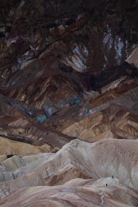 A dramatic landscape photograph of colorful, eroded badlands rock formations with striped patterns of brown, pink, and teal, featuring two tiny human figures for scale at the bottom.