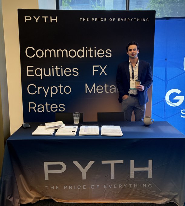 A man stands behind a table displaying some type of information. The text on the poster reads "Live from @neudatalab NYC. Stop by the Pyth booth to learn how market data providers, exchanges, and financial institutions are reaching the next generation of financial applications through a single integration". In this image, there is a person wearing a white shirt standing at a computer desk with a glass of water. The man appears to be presenting or talking about cryptocurrencies on the table in front of him, which indicates that he may have some knowledge and expertise related to cryptocurrency-related matters.