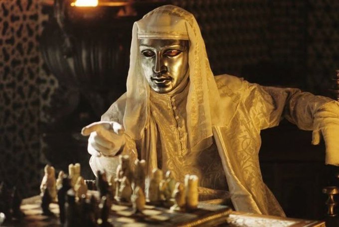 A masked figure in white robes and gloves plays chess in dim, candlelit surroundings, evoking a strategic mastermind plotting their next move. The image metaphorically illustrates the post's point about @Enter_Elysium executing a calculated, strategic maneuver by redirecting sequencer fees to builders—positioning itself as a cunning player making a powerful move on the crypto chessboard.