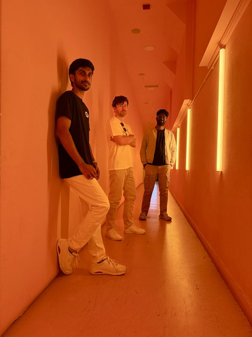 Three men pose along a narrow corridor bathed in warm orange light from vertical LED tubes, fitting the post's behind-the-scenes showcase of the 0G DevRel team during their time in Portugal.