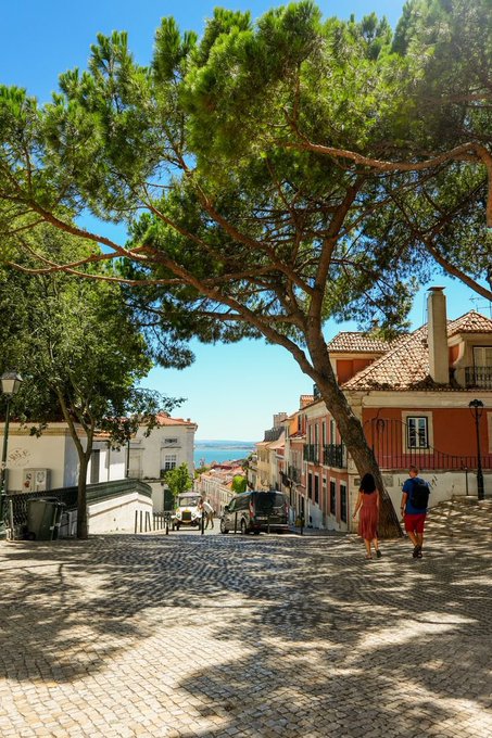 The image shows a cobblestone street with two people walking down it. There are also trees nearby that provide shade for the pedestrians. The scene appears to be set in a European town or city, as suggested by the presence of buildings along the street. A car is parked on the side of the road, and there's an advertisement posted on one of the buildings.
