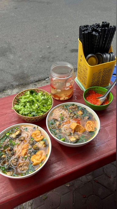 Two bowls of noodle soup with fried tofu, herbs, and greens sit on a red table alongside a bowl of cilantro, a small dish of chili sauce, a jar of pickled vegetables, and a yellow holder with chopsticks.