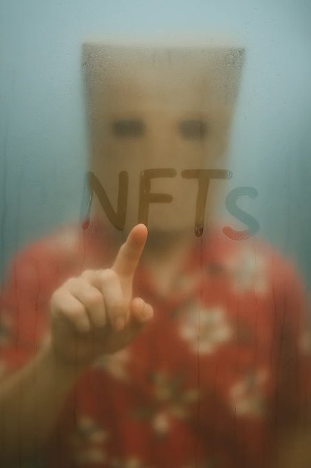A child stands behind foggy glass with their index finger raised, while the word "NFTS" is written across the condensation on the window. The hazy, obscured view through the glass mirrors the speculative and often unclear nature surrounding NFT investments.