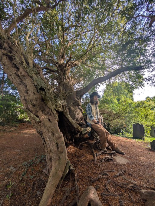A person wearing a hat, plaid shirt, and vest sitting against the gnarled trunk of a large tree in what appears to be a wooded area with gravestones visible in the background.