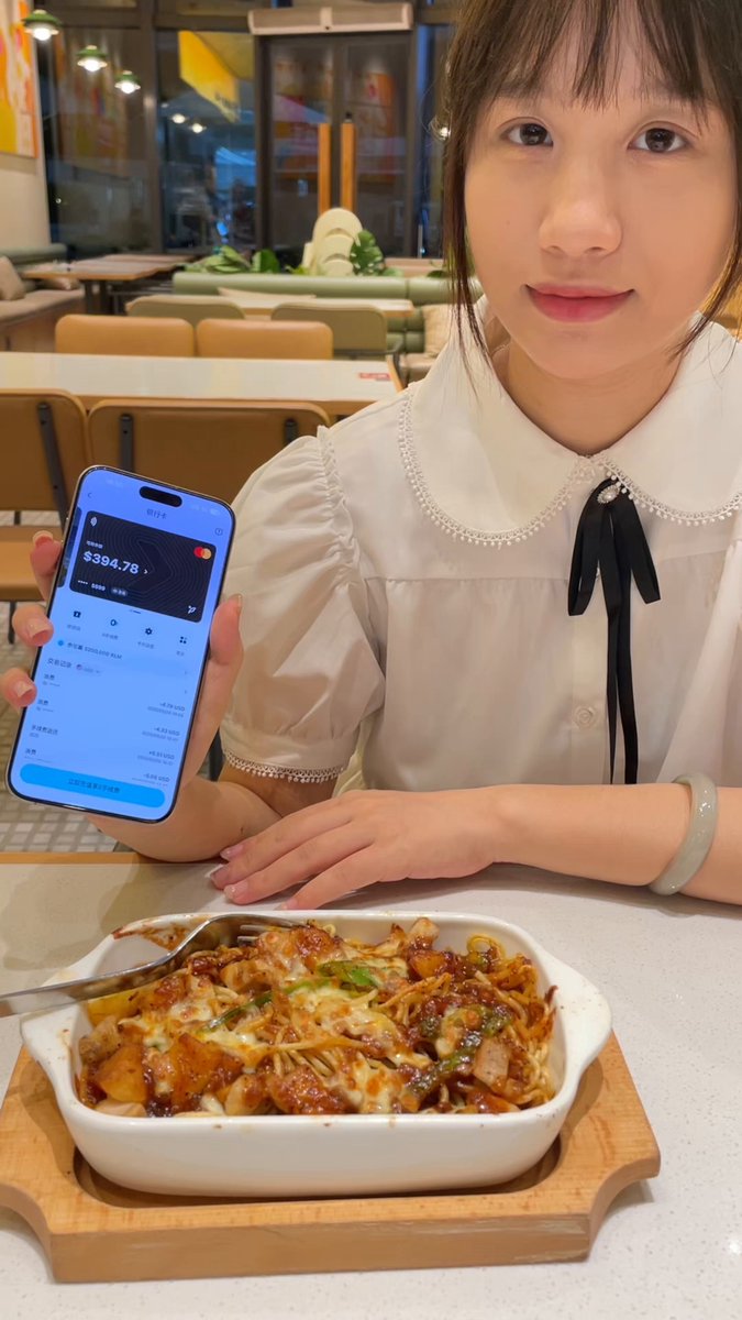 The image features a young woman sitting at a dining table with food, holding up her cell phone. She appears to be showing off something on her phone or possibly using an app related to cryptocurrency. The setting seems to be a restaurant, as there are chairs around the table and other people can be seen in the background.