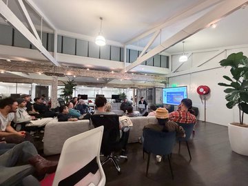 A diverse group gathers for a Starknet meetup in a bright, modern office space with exposed beams and industrial decor, with attendees seated and mingling as presentations are delivered.