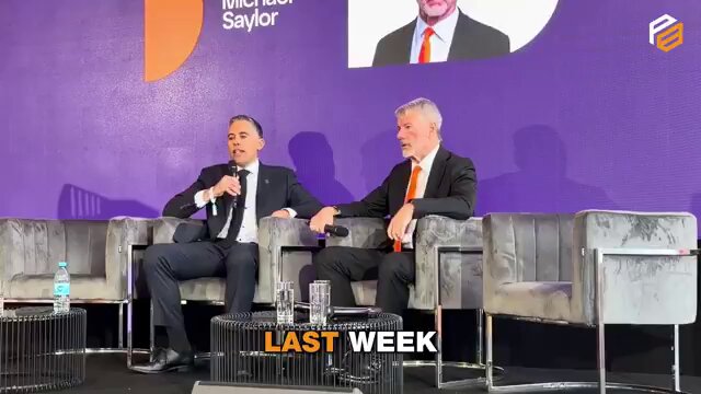 The image shows two men sitting next to each other, one wearing a suit and tie while the other has a tie with an orange logo. They are both holding microphones, suggesting they might be participating in a panel discussion or interview. The background features a purple wall with a picture of a man on it, as well as some chairs placed around them.

The post includes text that reads "LATEST: Michael Saylor responds to Strive CEO Matt Cole's joke about buying 32 Bitcoin". This indicates the image is related to an event or discussion where the two men are speaking and possibly discussing the topic of cryptocurrency.