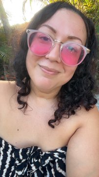 A selfie of a smiling woman with curly dark hair wearing pink-tinted round sunglasses and a black and white patterned top, taken outdoors.