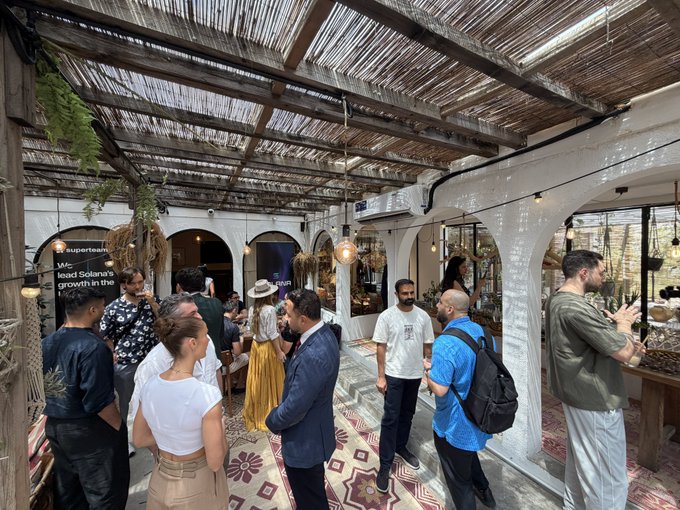 A photograph of a group of people mingling in a covered patio or courtyard setting, with arched white walls, a wooden slatted roof, string lights, and decorative floor tiles.