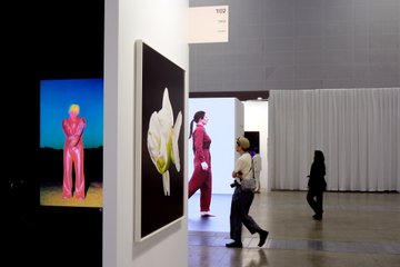 Art gallery interior with people walking past displays including a pink sculpture, abstract paintings, and a screen showing a figure.