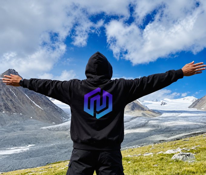 A person in a black hoodie with a purple and blue geometric logo stands triumphantly with arms outstretched, facing a stunning glacial mountain landscape under a partly cloudy sky—perfectly capturing the uplifting "gm ☀️⛰️" (good morning) vibe.