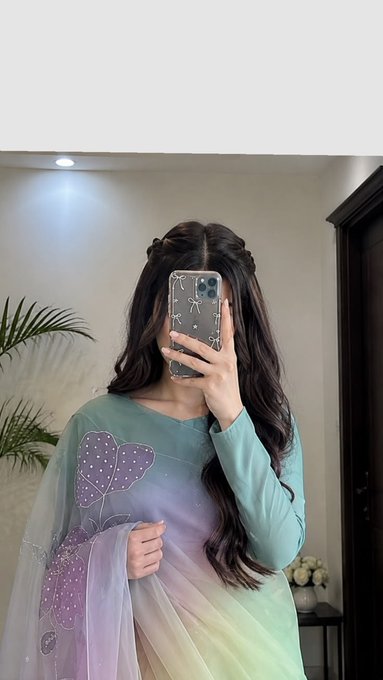 A mirror selfie of a person wearing a pastel-colored outfit with floral embroidery, taking the photo with a smartphone that obscures their face.