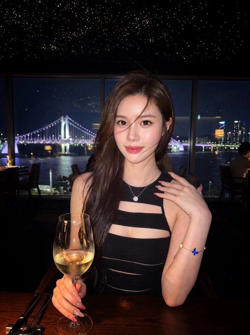 A young woman with long brown hair wearing a black cut-out dress holds a glass of white wine at a restaurant with large windows showing a nighttime cityscape and illuminated bridge.