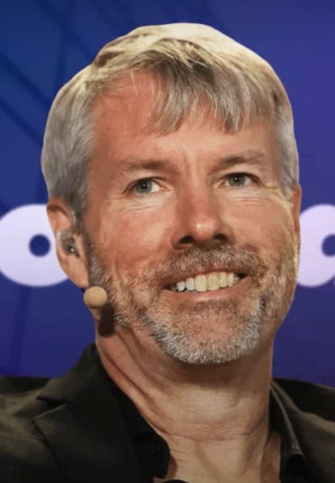 A close-up headshot of a smiling older man with grey hair and a beard, wearing a black shirt and an earpiece microphone, against a blue background.