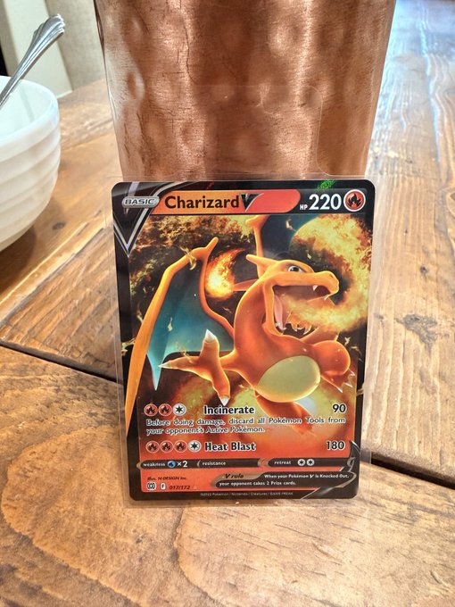 A holographic Charizard V Pokémon trading card (HP 220) with 'Incinerate' and 'Heat Blast' attacks rests on a wooden table next to a white bowl.