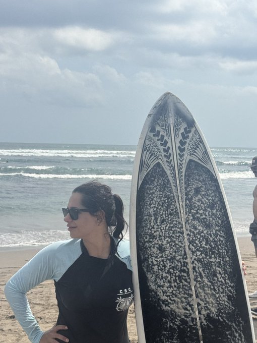 A woman wearing sunglasses and a black and light blue rashguard (with 'CSC' logo) standing on a beach next to a black surfboard with white decorative patterns.