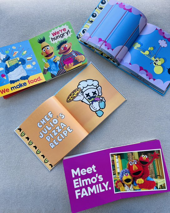 An overhead view of four Sesame Street-themed children's books laid on fabric, featuring titles and characters like Bert and Ernie with 'We're hungry!', Cookie Monster with 'Chef Julio's Pizza Recipe', and Elmo with 'Meet Elmo's FAMILY.'