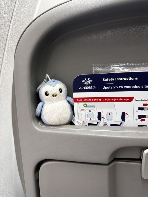 A small plush penguin sits in an airplane seat pocket next to an AirSERBIA safety instructions card labeled 'Take-off and Landing | Poletanje i sletanje'.