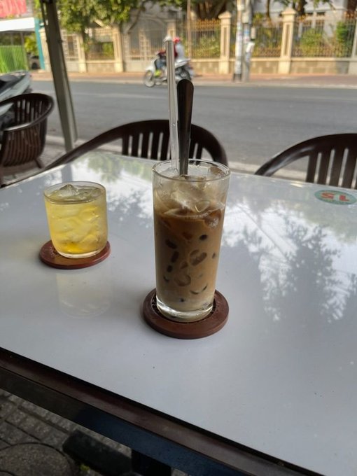 Two iced beverages, including what appears to be an iced coffee and a yellow drink, sit on a white cafe table overlooking a street with a motorbike passing by.