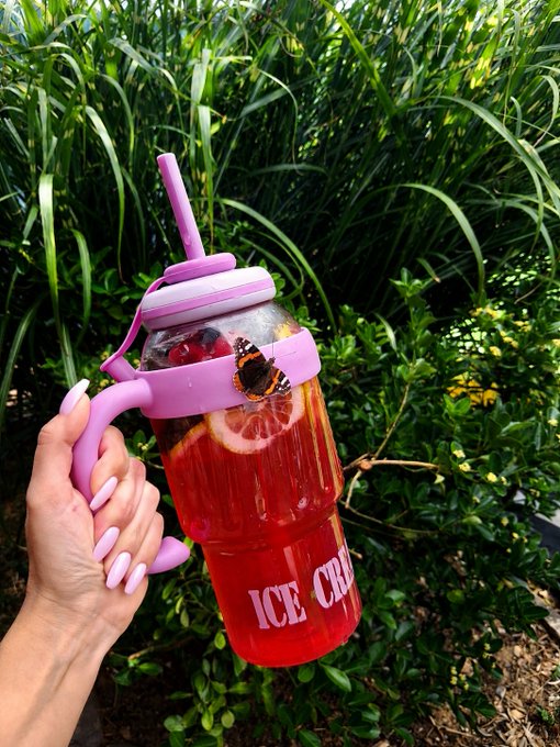 A hand with lavender-painted nails holding a pink and purple 'ICE CREAM' branded tumbler filled with a red fruit drink garnished with lemon, berries, and a butterfly decoration, set against green foliage.