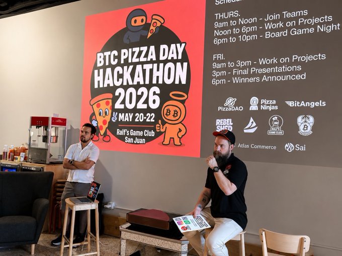 Two men at the BTC Pizza Day Hackathon 2026, held May 20-22 at Ralf's Game Club in San Juan, with event schedule and sponsors displayed on a wall.